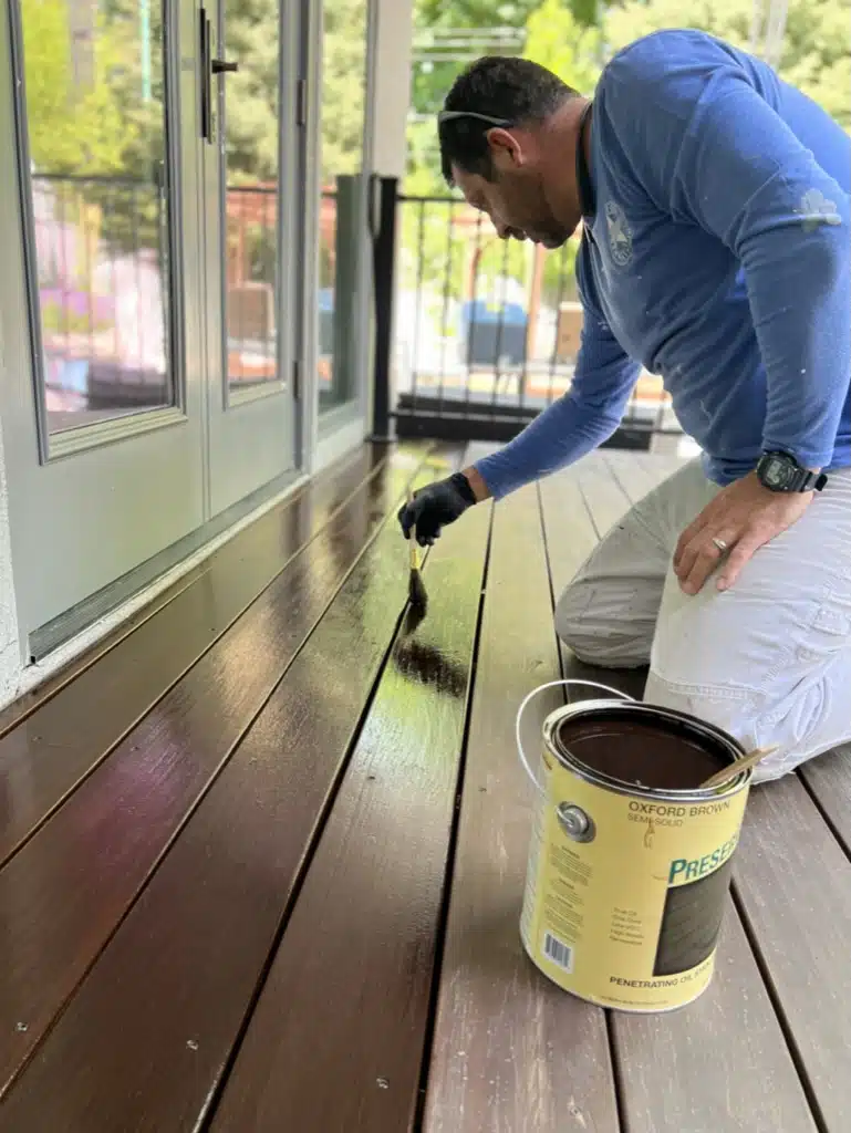 A man paints a wooden deck with a can of paint, representing American Town Painting's deck staining services