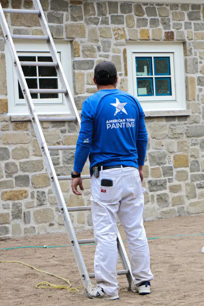 A man using a ladder to paint a house, showcasing the work of American Town Painting.