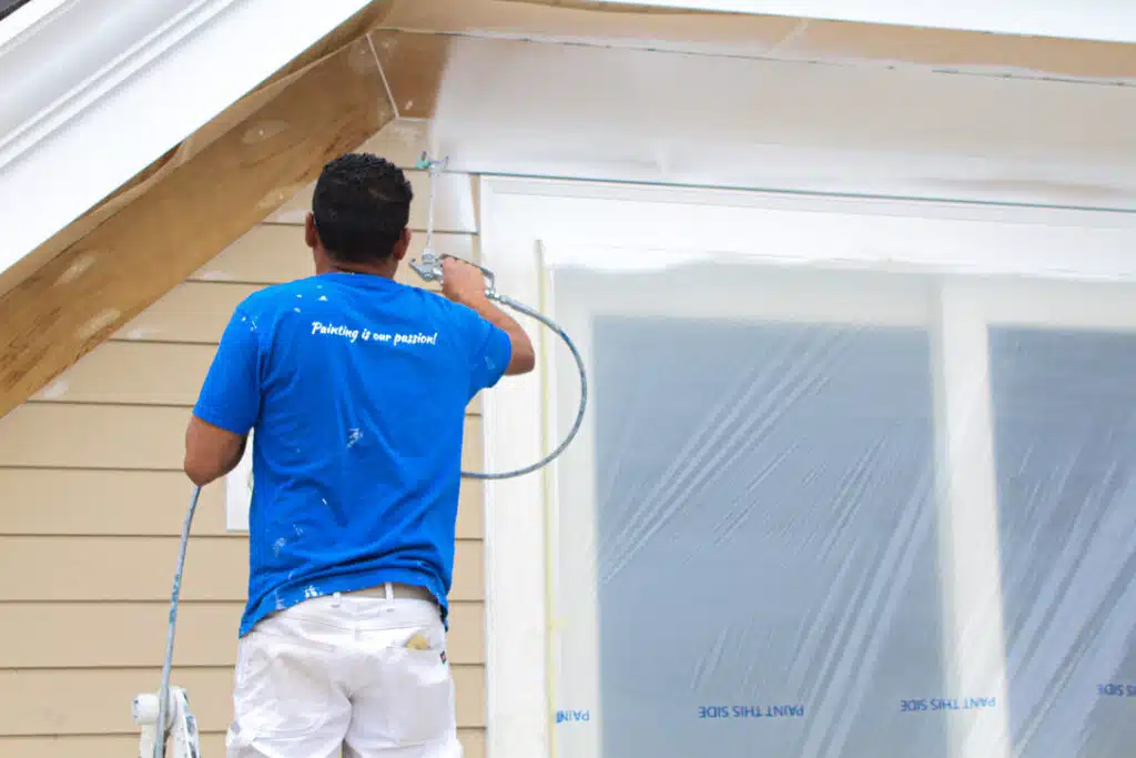 A man in a blue shirt paints a house, showcasing the work of American Town Painting.