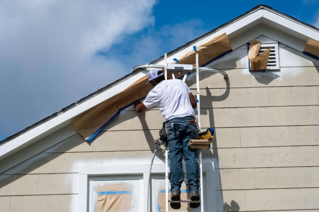 A contractor in a white shirt and jeans works from a ladder on a house roof repairing siding