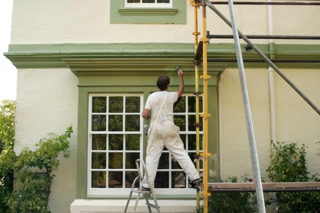 A contractor in a white shirt and jeans works from a ladder on a house roof repairing siding